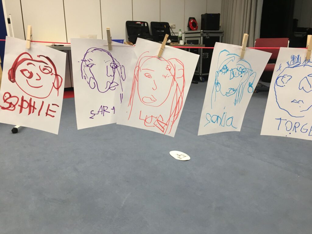 Fünf bunt gezeichnete Selbstporträts auf Papier, beschriftet mit den Namen Sophie, Sara, Lea, Sonja und Torge, hängen mit Holzwäscheklammern an einer roten Schnur im Seminarraum.