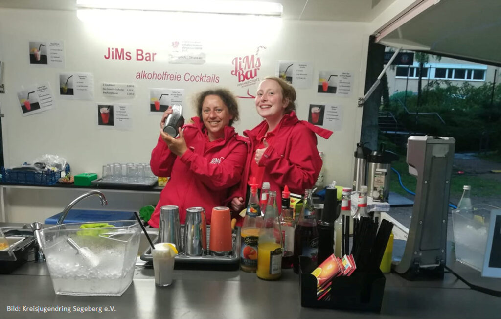 Mitarbeiterinnen bereiten alkoholfreie Cocktails an der mobilen 'JiMs Bar' des Kreisjugendrings Segeberg e.V. zu. Eine Mitarbeiterin hält den Shaker in den Händen, während die andere Mitarbeiterin freundlich mit ihrem Daumen nach oben zeigend in die Kamera lächelt.