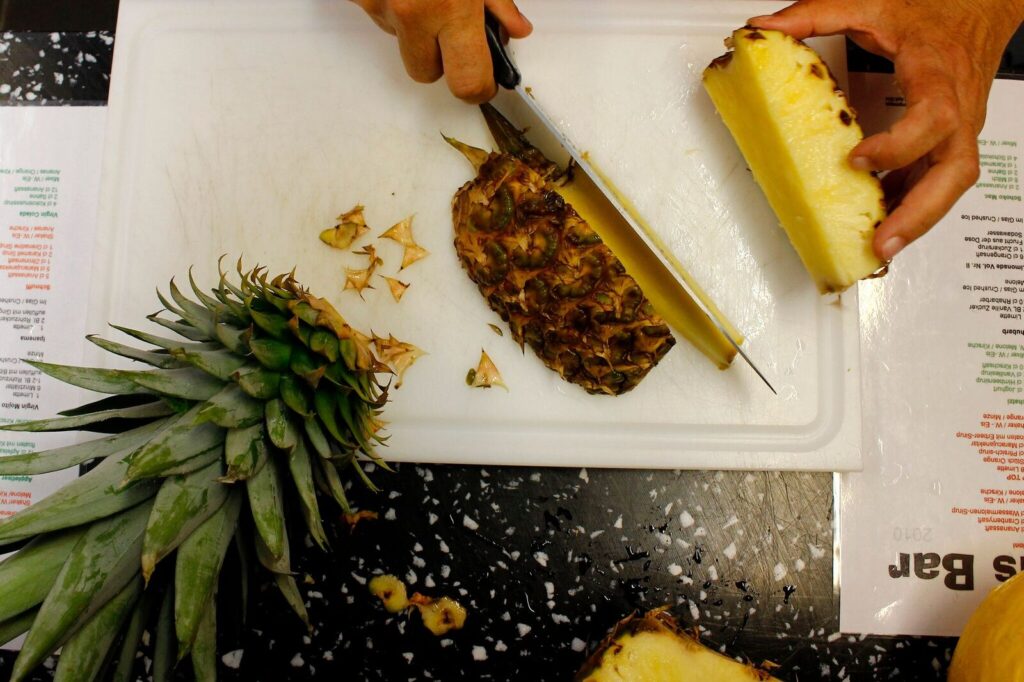 Vogelperspektive: Eine Person schneidet mit einem Messer eine frische Ananas auf einem weißen Schneidebrett zu