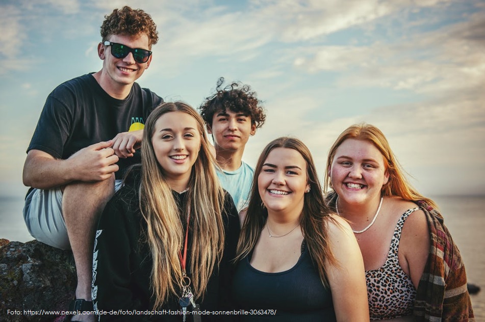 Eine Gruppe von fünf fröhlichen jungen Menschen sitzt und steht eng beieinander im Freien am Wasser und lächelt in die Kamera.