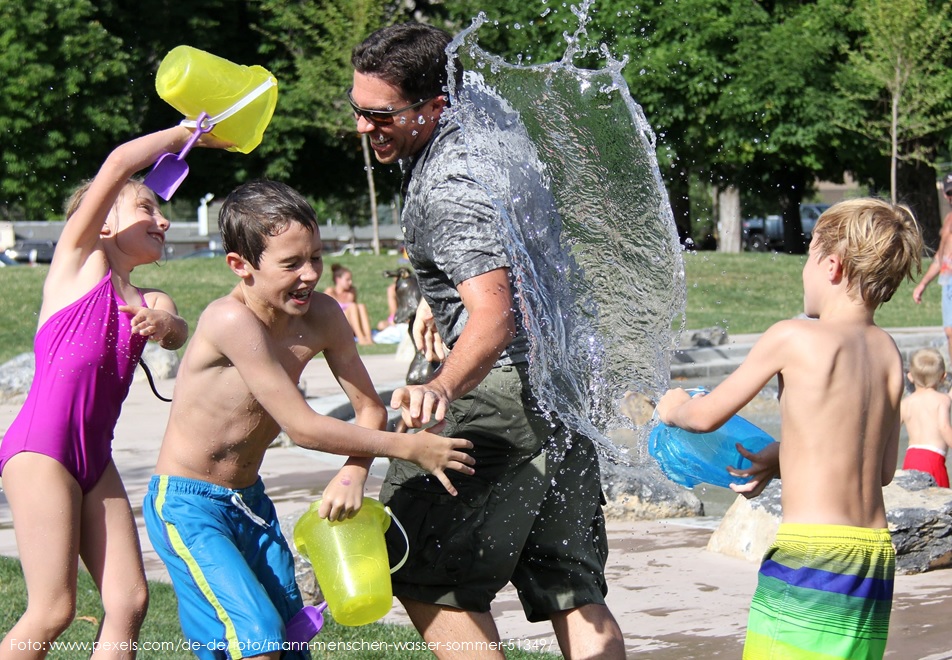 Ein lachender Mann und drei Kinder in Badebekleidung liefern sich im Freien eine ausgelassene Wasserschlacht und schütten Wasser aus bunten Plastikeimern aufeinander.