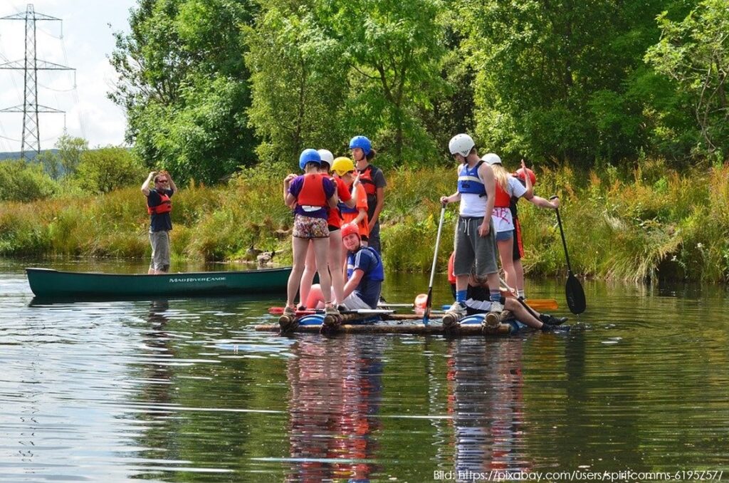 Eine Gruppe von neun Jugendlichen mit Schutzhelmen und Schwimmwesten steht gemeinsam auf einem selbstgebauten Floß auf einem ruhigen Fluss. Im Hintergrund ist eine grüne Naturkulisse zu sehen.